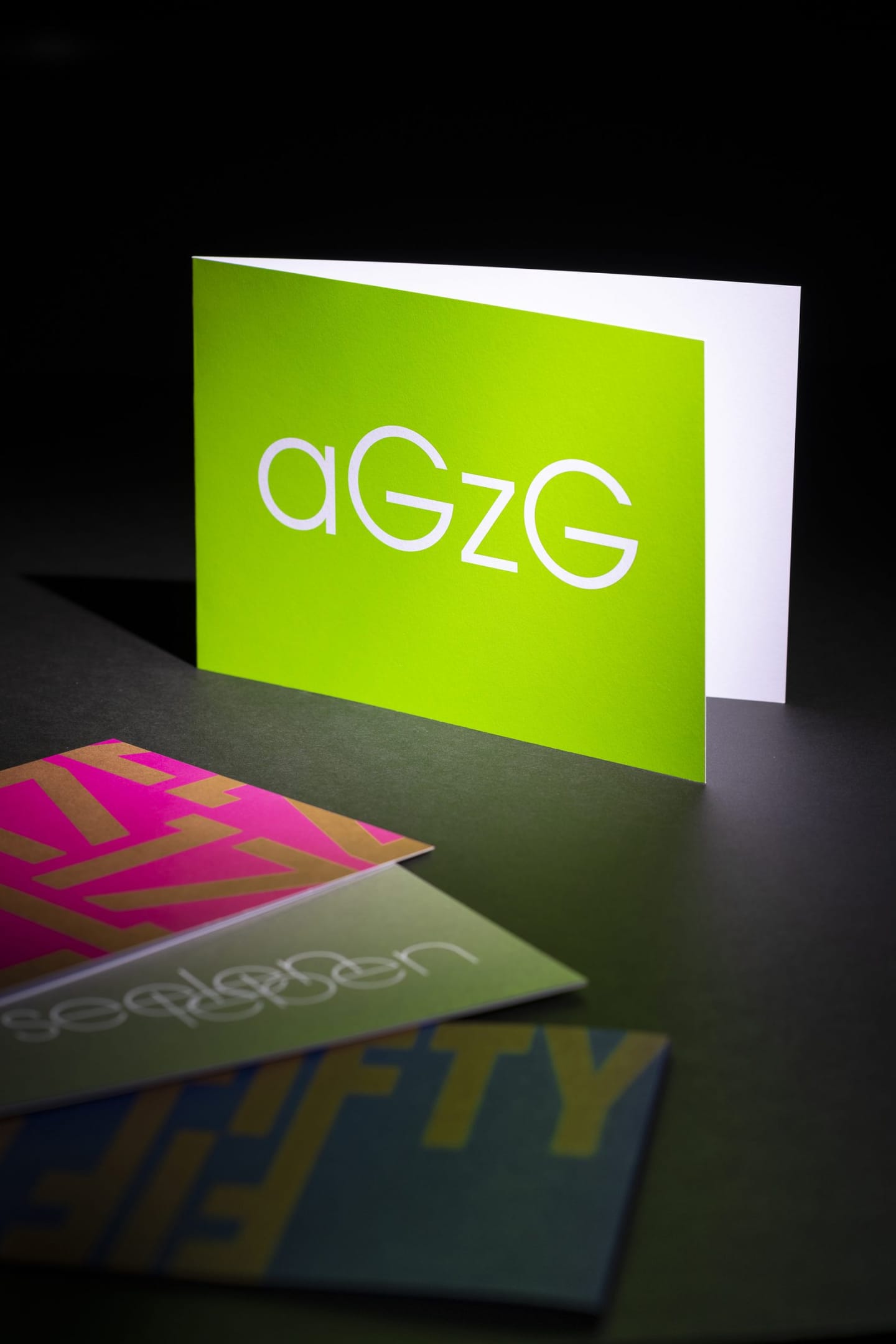 Eine aufgestellte grüne Klappkarte mit dem Schriftzug aGzG, davor liegen weitere Karten in Pink, Grün und Blau.